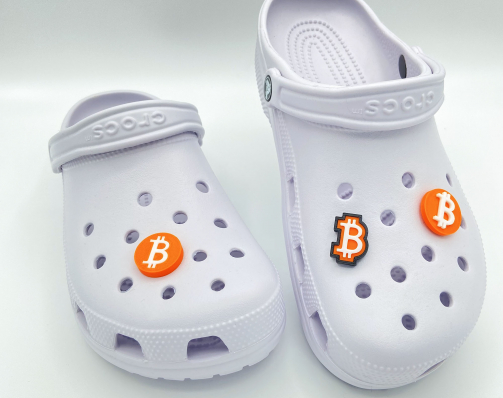 A pair of white clogs featuring a collection of three different 3D printed orange Bitcoin shoe charms.